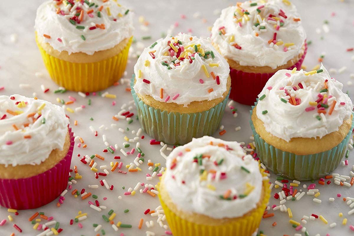 Several frosted cupcakes with colorful sprinkles in vibrant paper liners scattered on a marble surface.