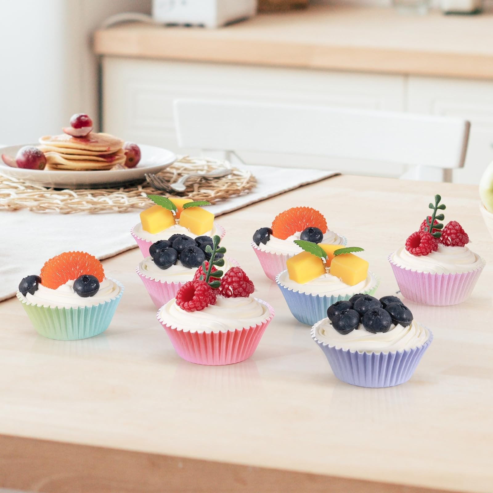 Assortment of decorated cupcakes in pastel-colored liners on a table, with a stack of pancakes in the background.