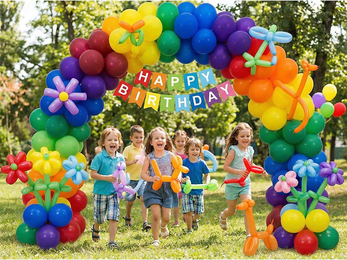 Children running through a rainbow balloon arch with a 'Happy Birthday' banner in a park setting.