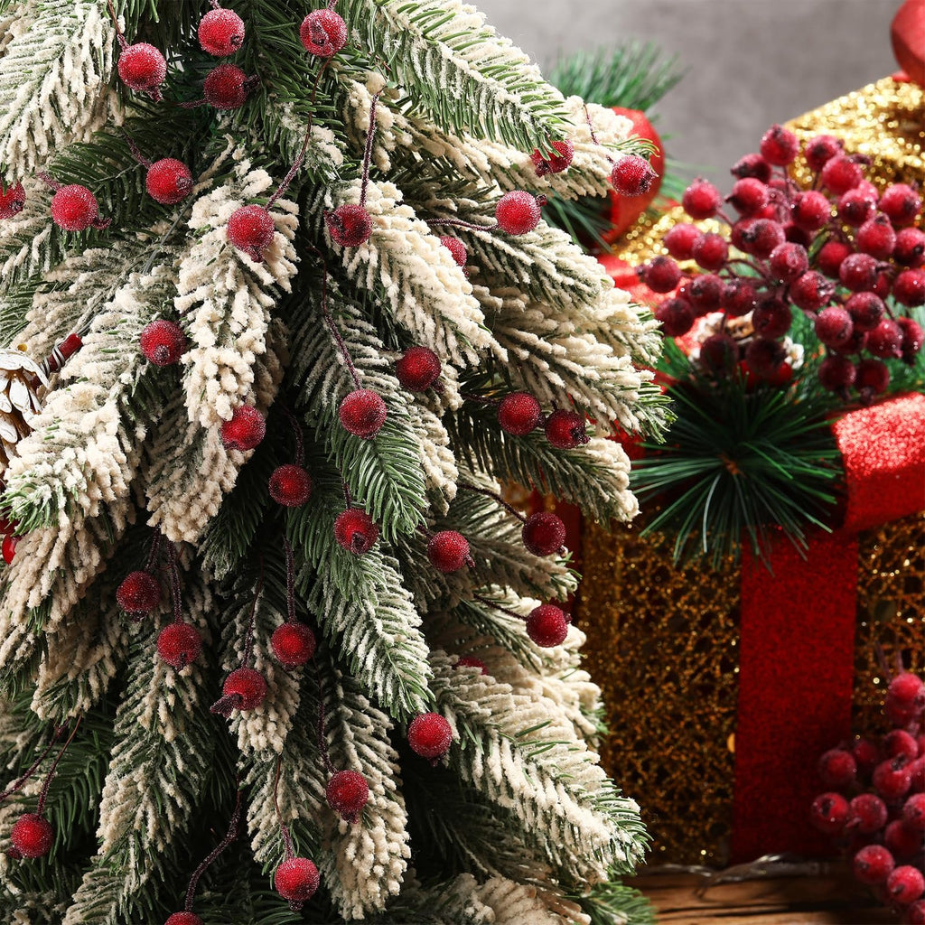 Frosted artificial holly berry stems with dark red berries on a flocked Christmas tree branch, with festive gift boxes in the background.