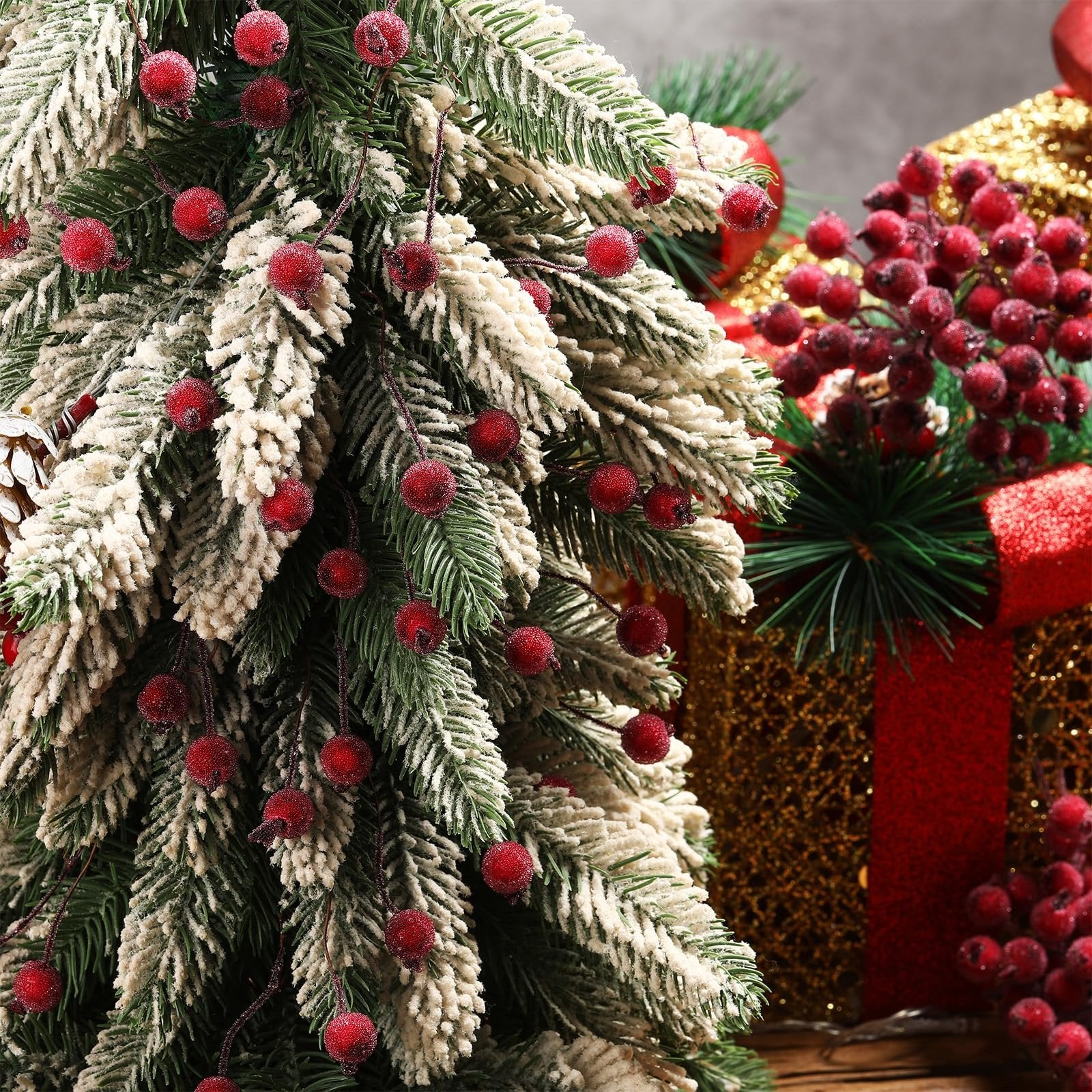 Frosted artificial holly berry stems with dark red berries on a flocked Christmas tree branch, with festive gift boxes in the background.