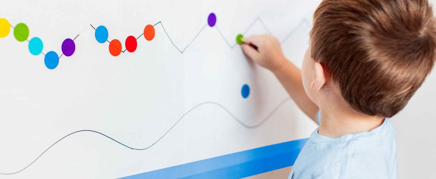 Child's hand placing a green dot sticker on a graph drawn on a whiteboard with colorful dot stickers
