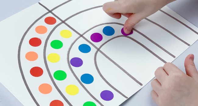 Child's hands placing colorful dot stickers onto a rainbow drawing on white paper.
