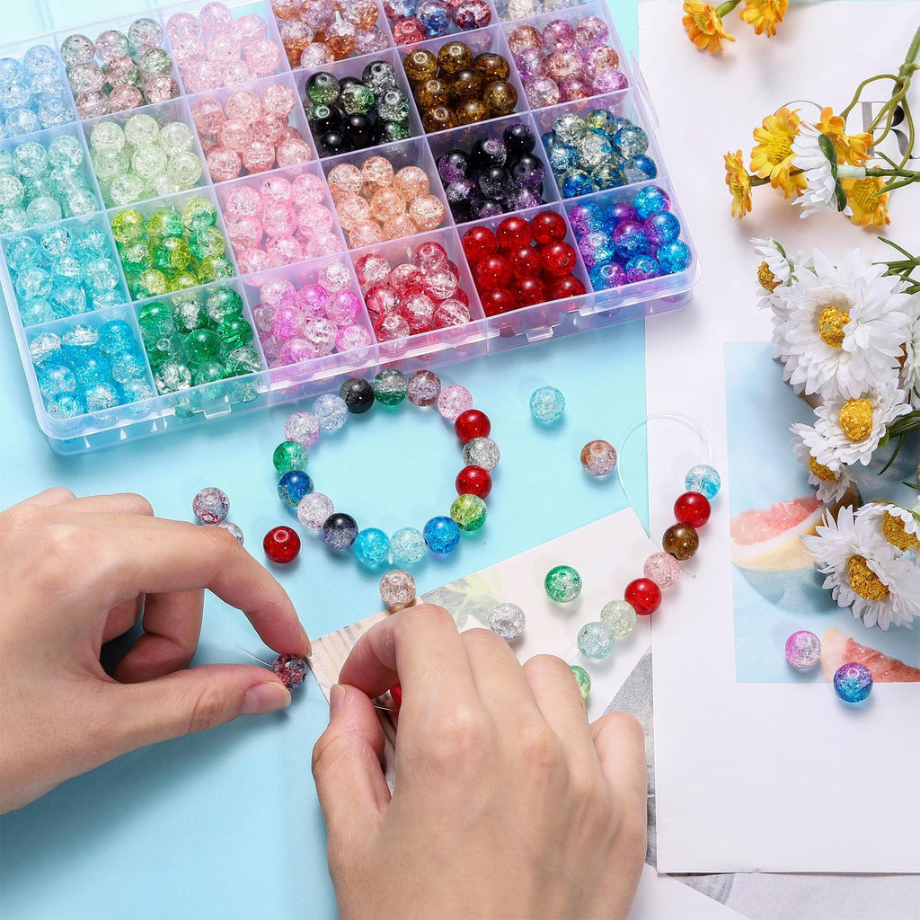 Hands stringing colorful crackle glass beads from a divided storage box, with a bracelet and loose beads scattered around.