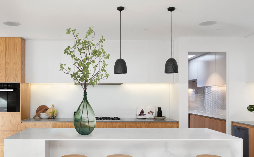 Artificial ficus greenery stems in a large green glass vase on a kitchen island, with modern cabinetry and pendant lights in the background.
