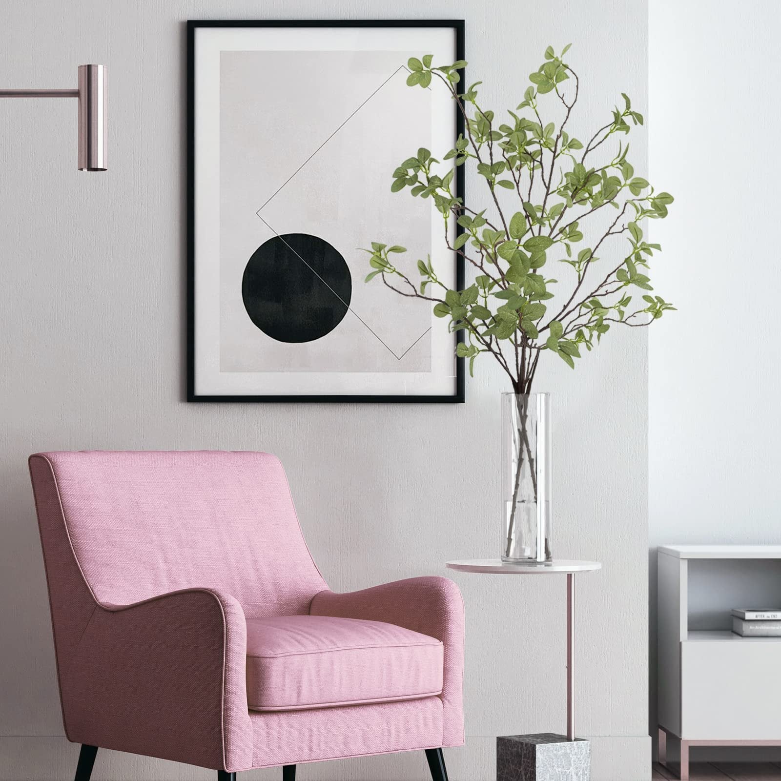 Artificial ficus twig greenery stems in a glass vase on a side table next to a pink armchair and abstract art.