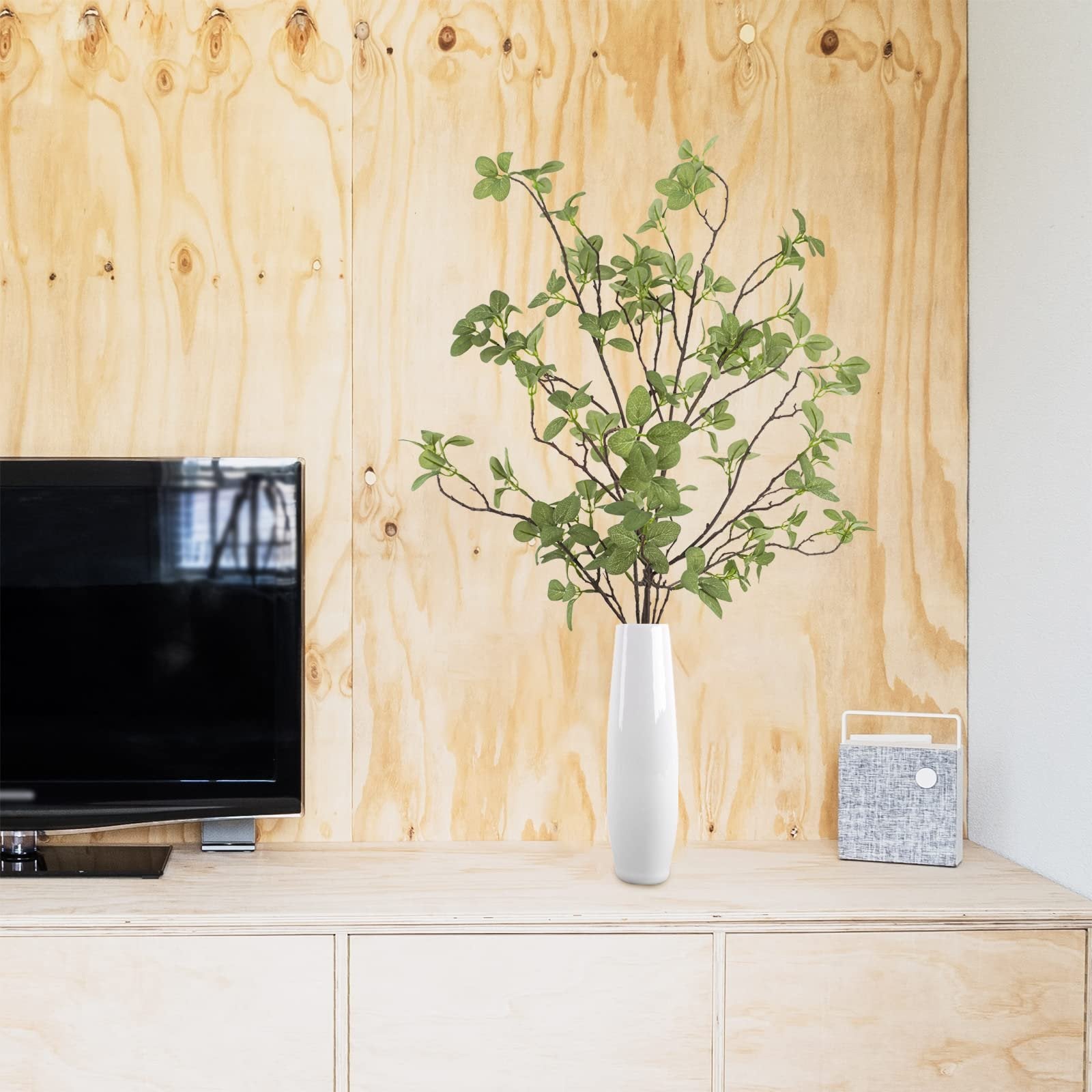 Artificial ficus twig greenery stems in a white vase on a wooden cabinet next to a television and a decorative speaker.