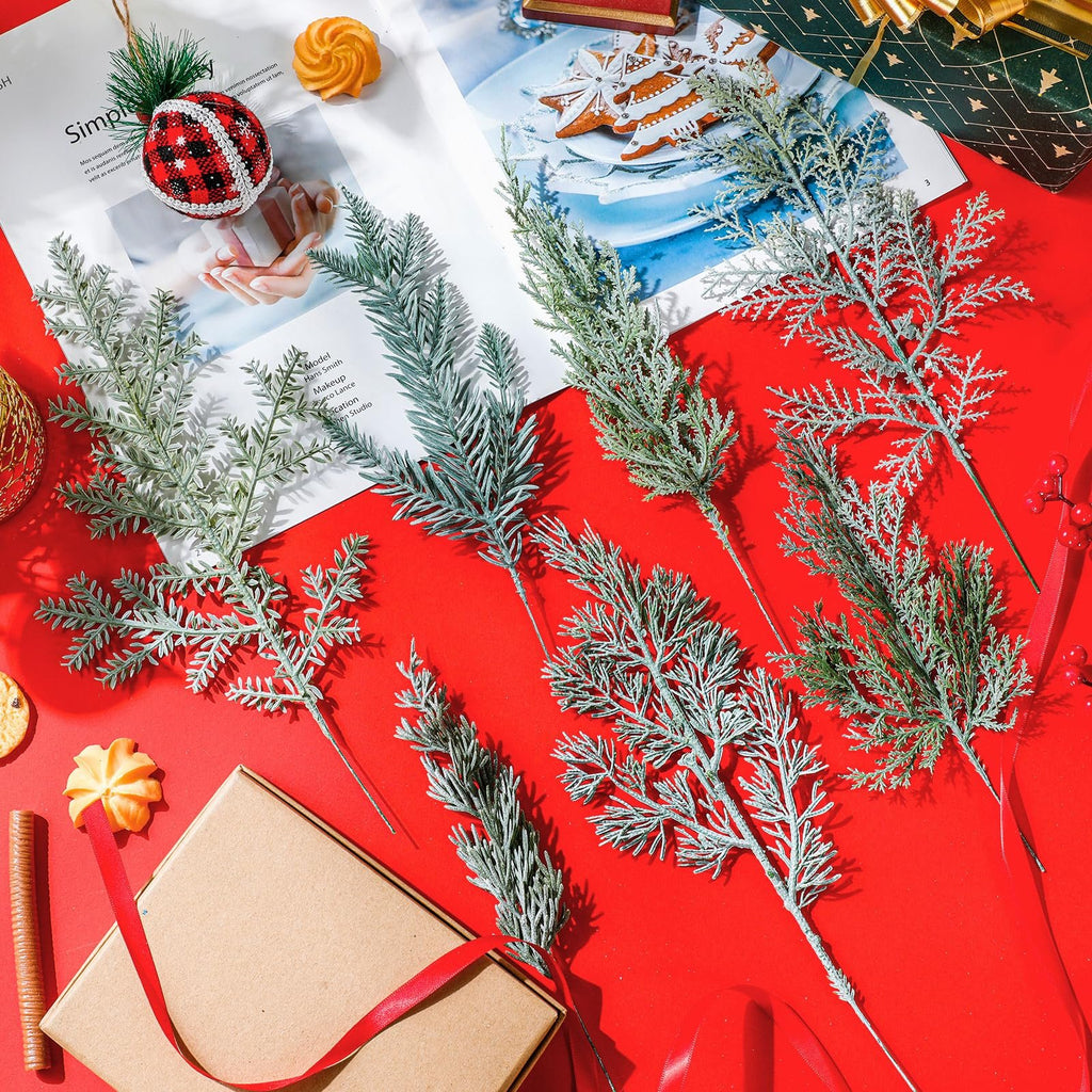 Seven artificial pine branches with flocked and green foliage arranged on a red surface with a gift box and cookies.