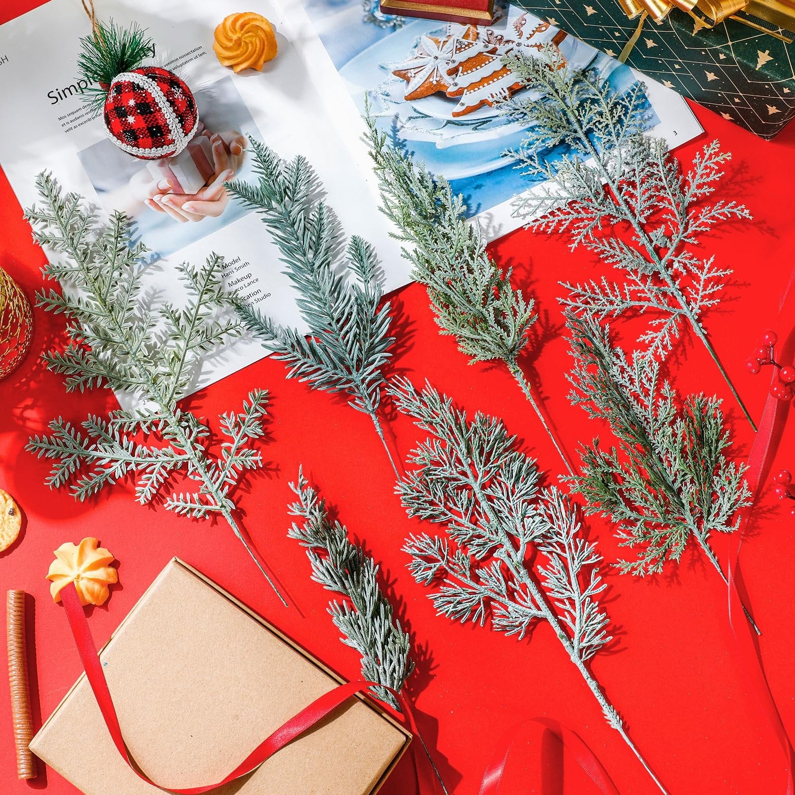 Seven artificial pine branches with flocked and green foliage arranged on a red surface with a gift box and cookies.