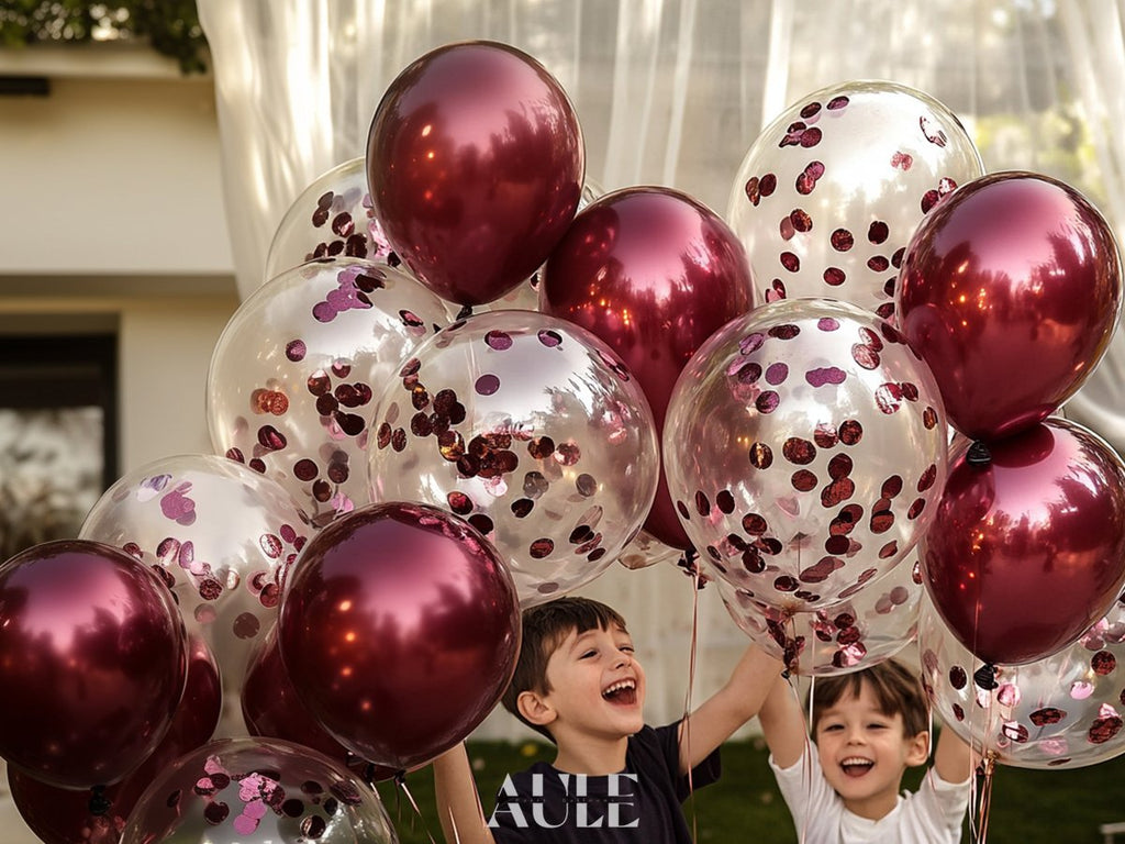 Two happy children holding a bouquet of burgundy and confetti balloons outdoors.
