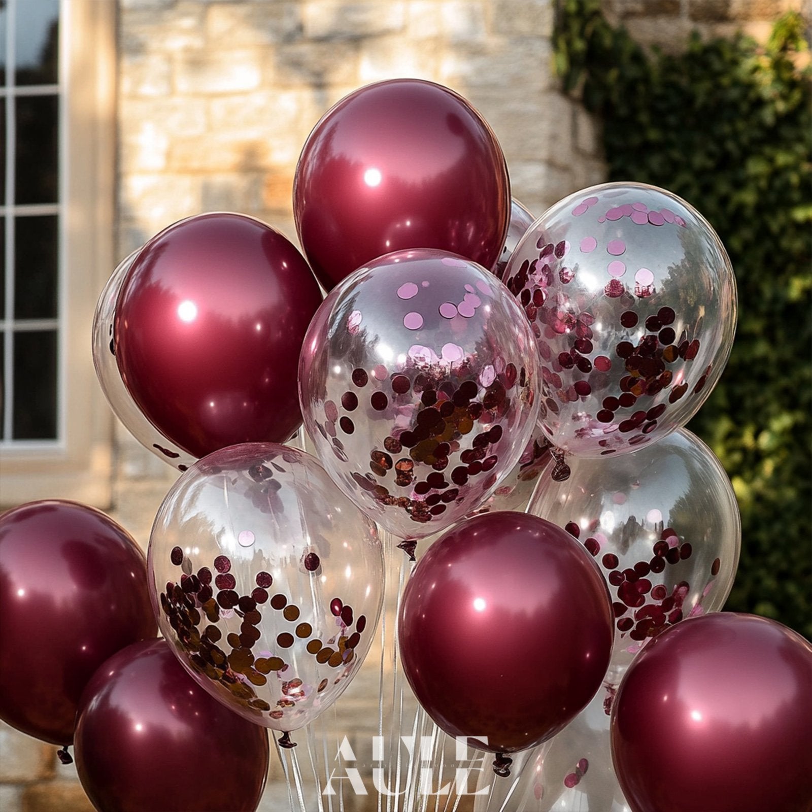 Burgundy and confetti balloons arranged in a bouquet outdoors, with a brick building in the background.