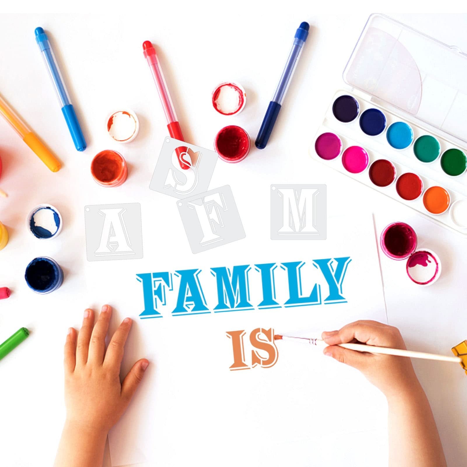 Child's hands painting the word FAMILY IS using stencils and watercolors on white paper, with art supplies scattered around.