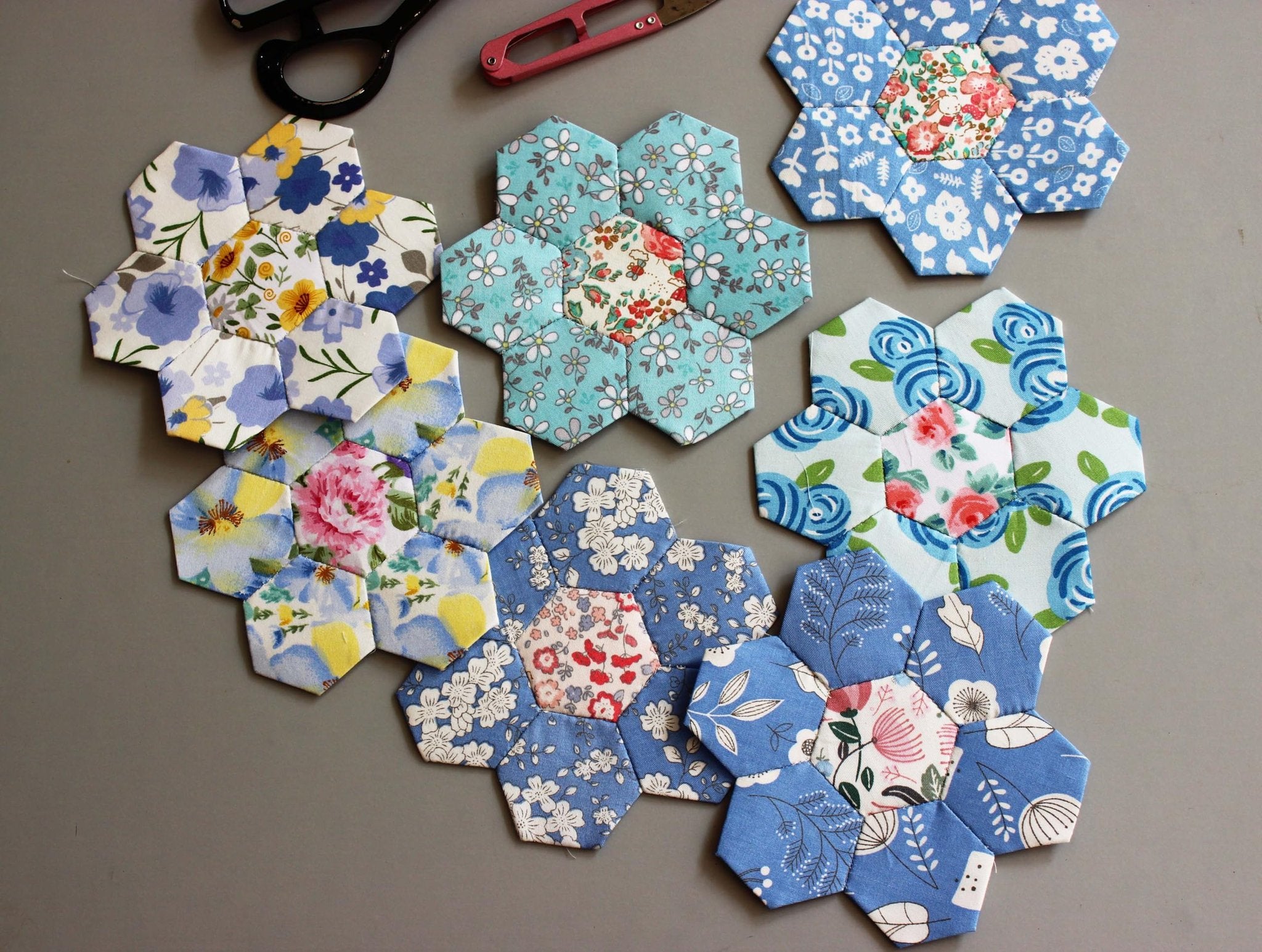 Hexagonal fabric pieces with floral patterns arranged on a gray surface, with scissors and a seam ripper in the background.