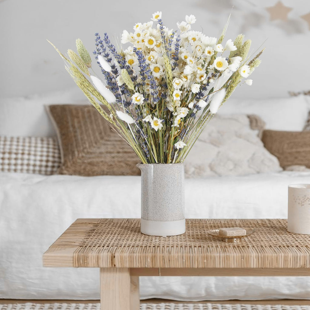 Dried flower bouquet with lavender, daisies, and wheat in a speckled vase on a woven bench.