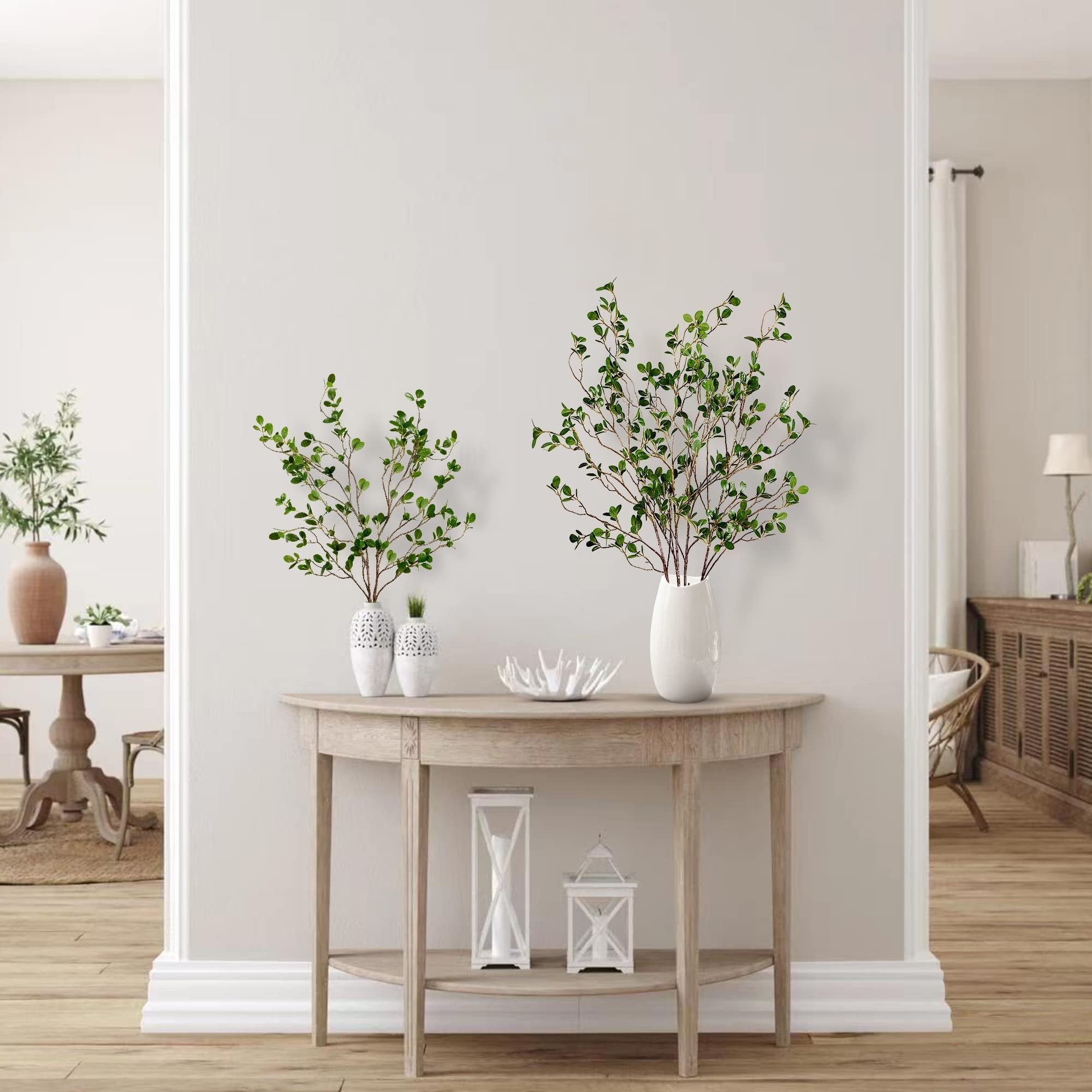 Two arrangements of artificial green ficus branches in white vases on a wooden console table.