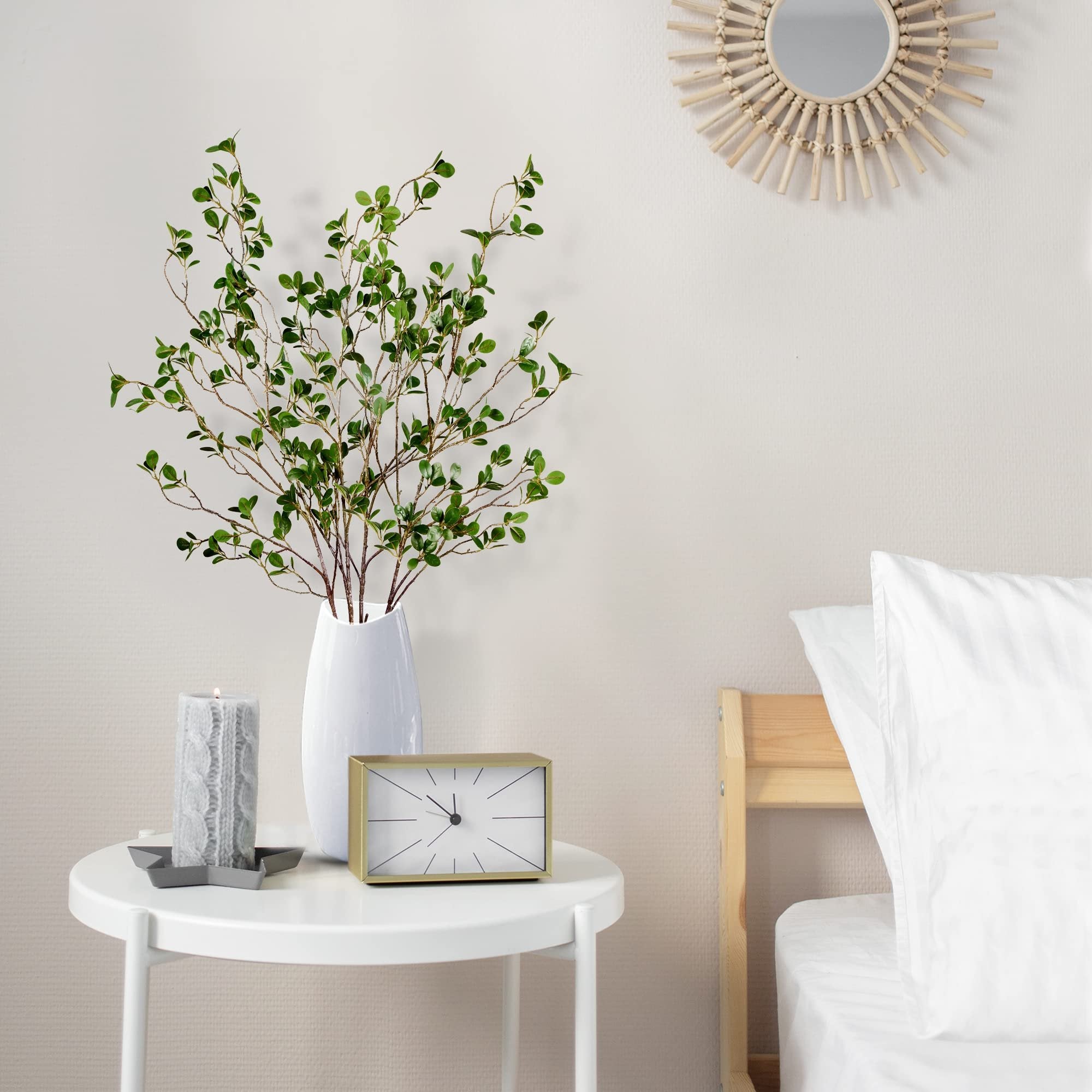 Artificial green ficus branches in a white vase on a side table next to a bed, with a candle and clock.