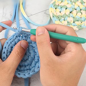 Close-up of hands crocheting with a blue yarn and a teal ergonomic crochet hook with 5.5mm size marking.