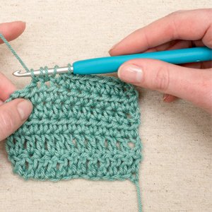 Hands crocheting with a teal ergonomic hook and green yarn on a textured surface.