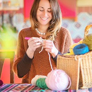 Woman smiling while crocheting with yarn and a pink crochet hook, basket of yarn nearby.