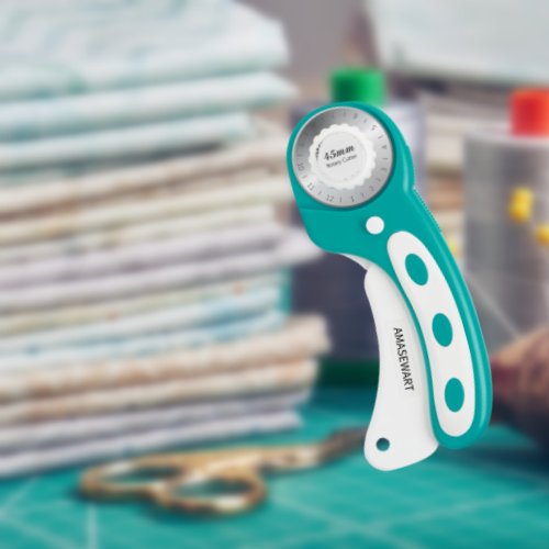 Turquoise rotary cutter with white handle, showing 45mm size on blade guard, with fabric stack and scissors in background.