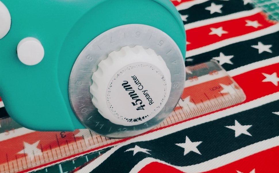 Close-up of a teal rotary cutter with a white dial on striped fabric with white stars. A ruler is visible underneath.
