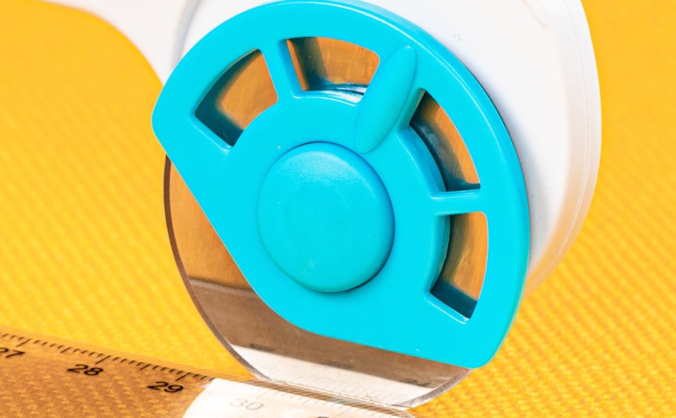 Close-up of a 45mm rotary cutter with a blue safety guard and metal blade, positioned over a ruler on a yellow textured surface.
