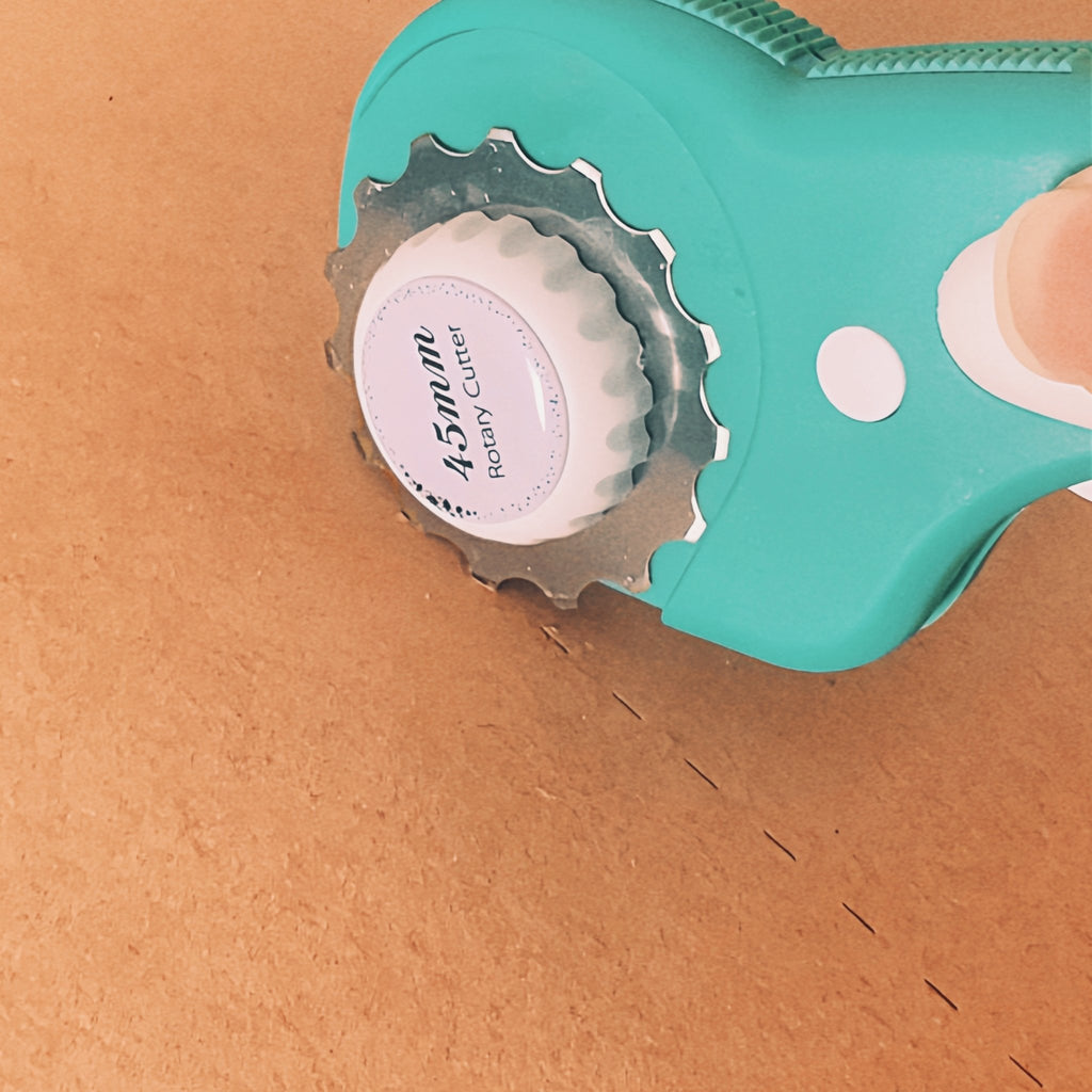 Close-up of a teal rotary cutter with a pink label cutting a dashed line on brown material.