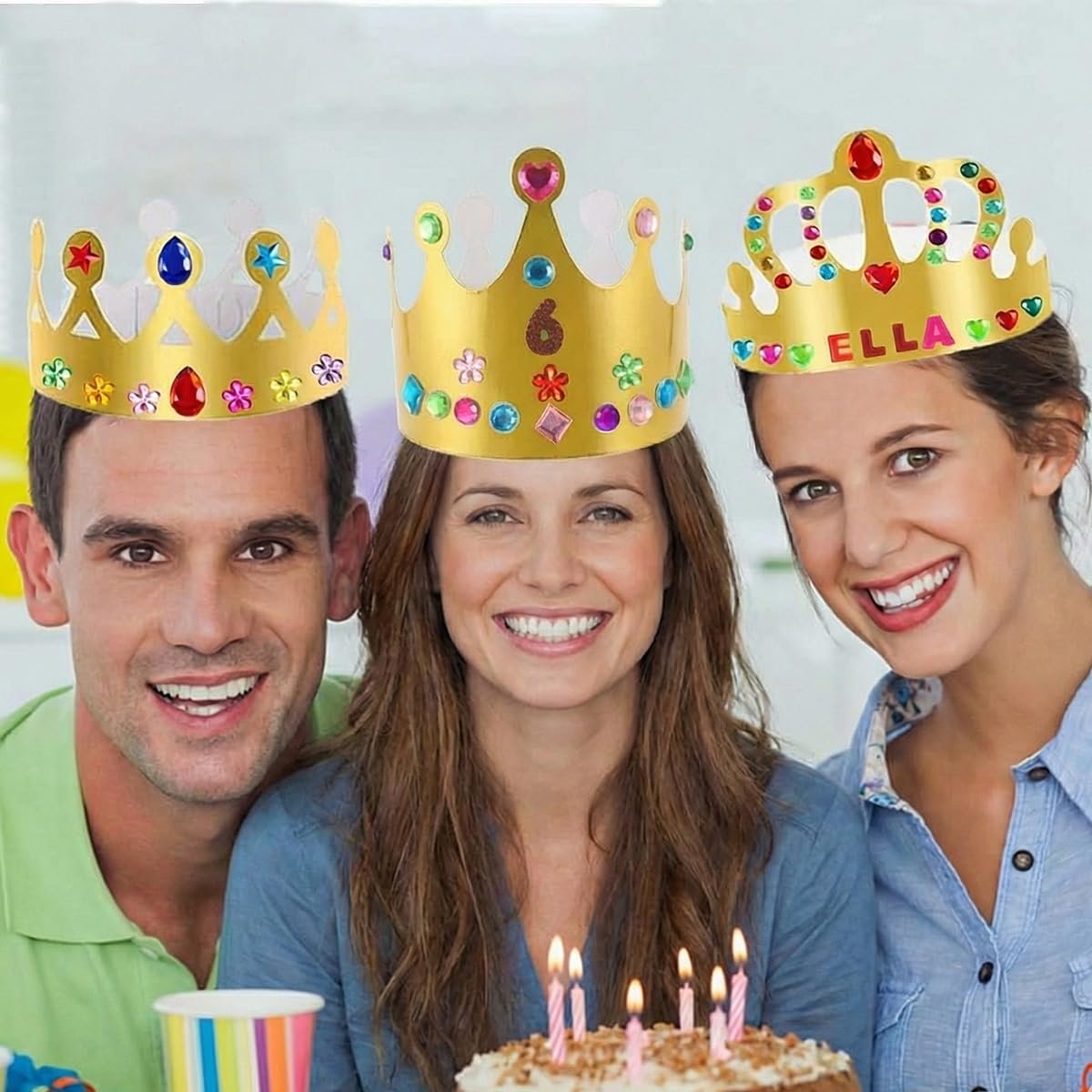 Three smiling people wearing decorative gold paper crowns with colorful jewels and stickers, celebrating with a birthday cake.