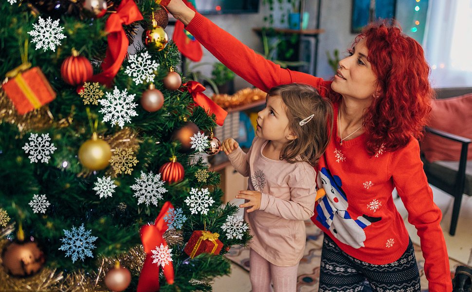 Woman and child decorating a Christmas tree with white glitter snowflake ornaments and red baubles.