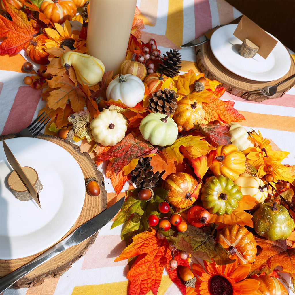 Fall harvest decor set with artificial pumpkins, gourds, pinecones, acorns, and maple leaves arranged on a striped tablecloth.