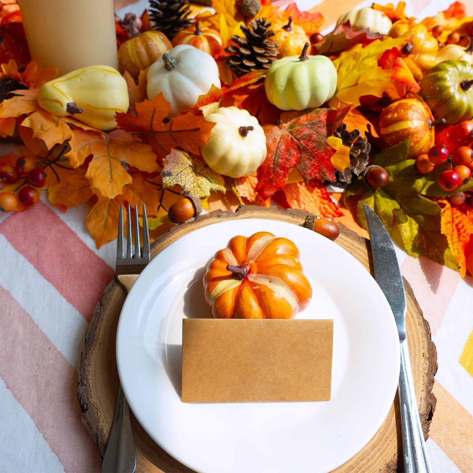 Table setting with a white plate, a small orange pumpkin, and a kraft paper card, surrounded by artificial fall leaves, gourds, acorns, and pinecones.