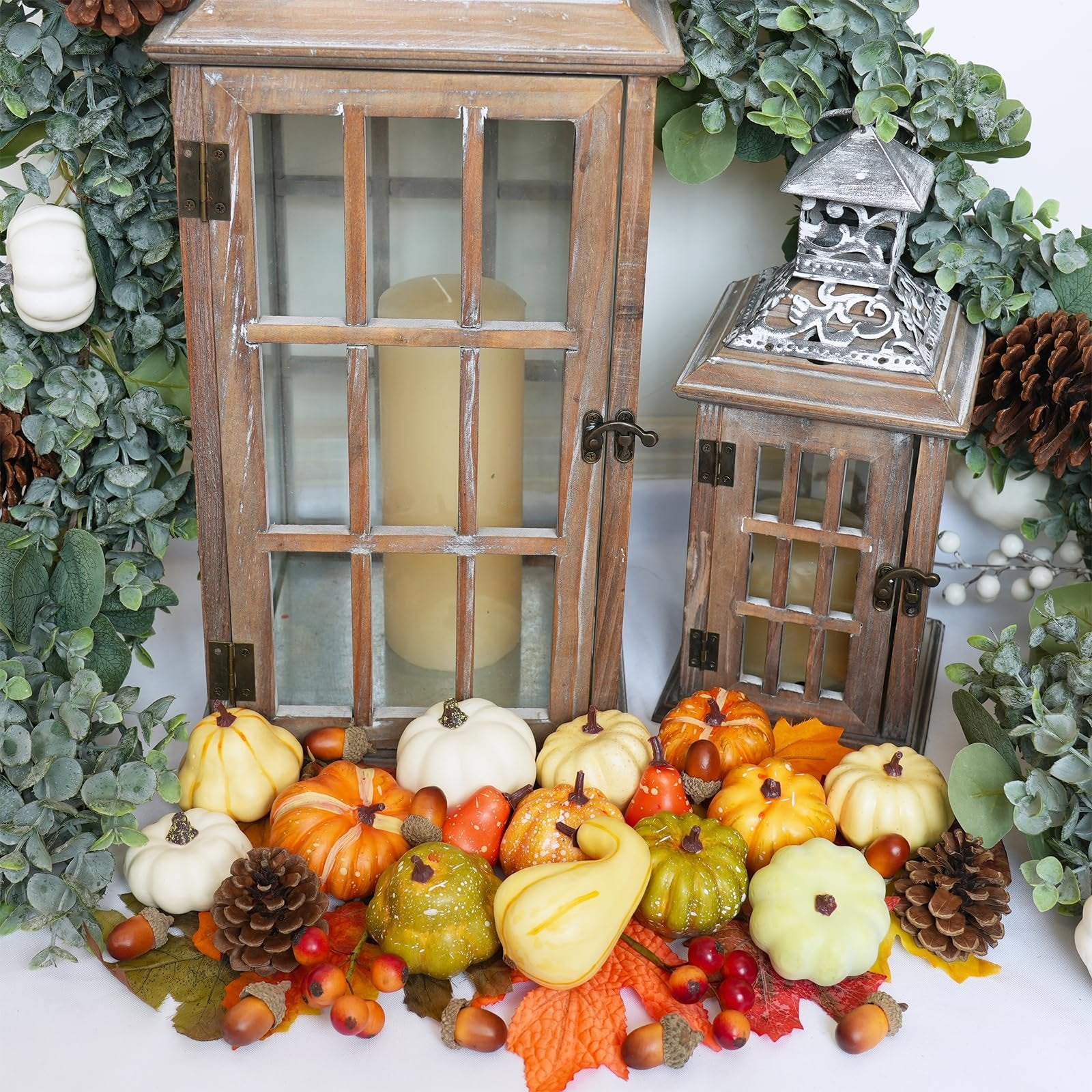 Assortment of artificial fall harvest decor including pumpkins, gourds, acorns, pinecones, and maple leaves arranged around wooden lanterns.