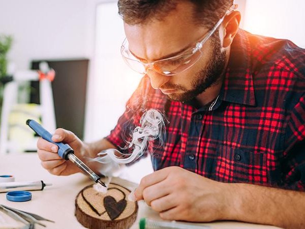 Man using a wood burning tool to create a heart design on a wooden slice, smoke rising from the tool.