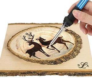 Hand holding a wood burning pen, creating a deer design on a wood slice.