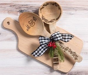 Two wooden spoons with pyrography designs and initials M & D, tied with a bow and greenery, resting on a wooden cutting board.