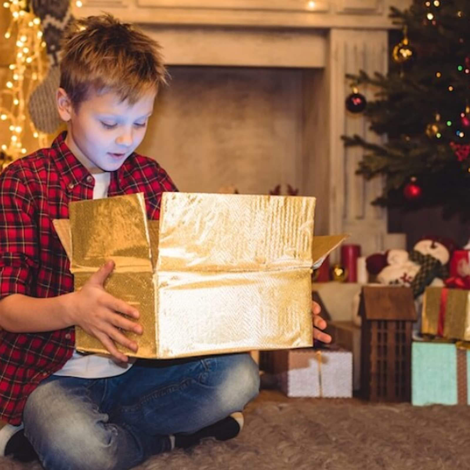 Boy opening a golden gift box near a Christmas tree and fireplace, with presents around.