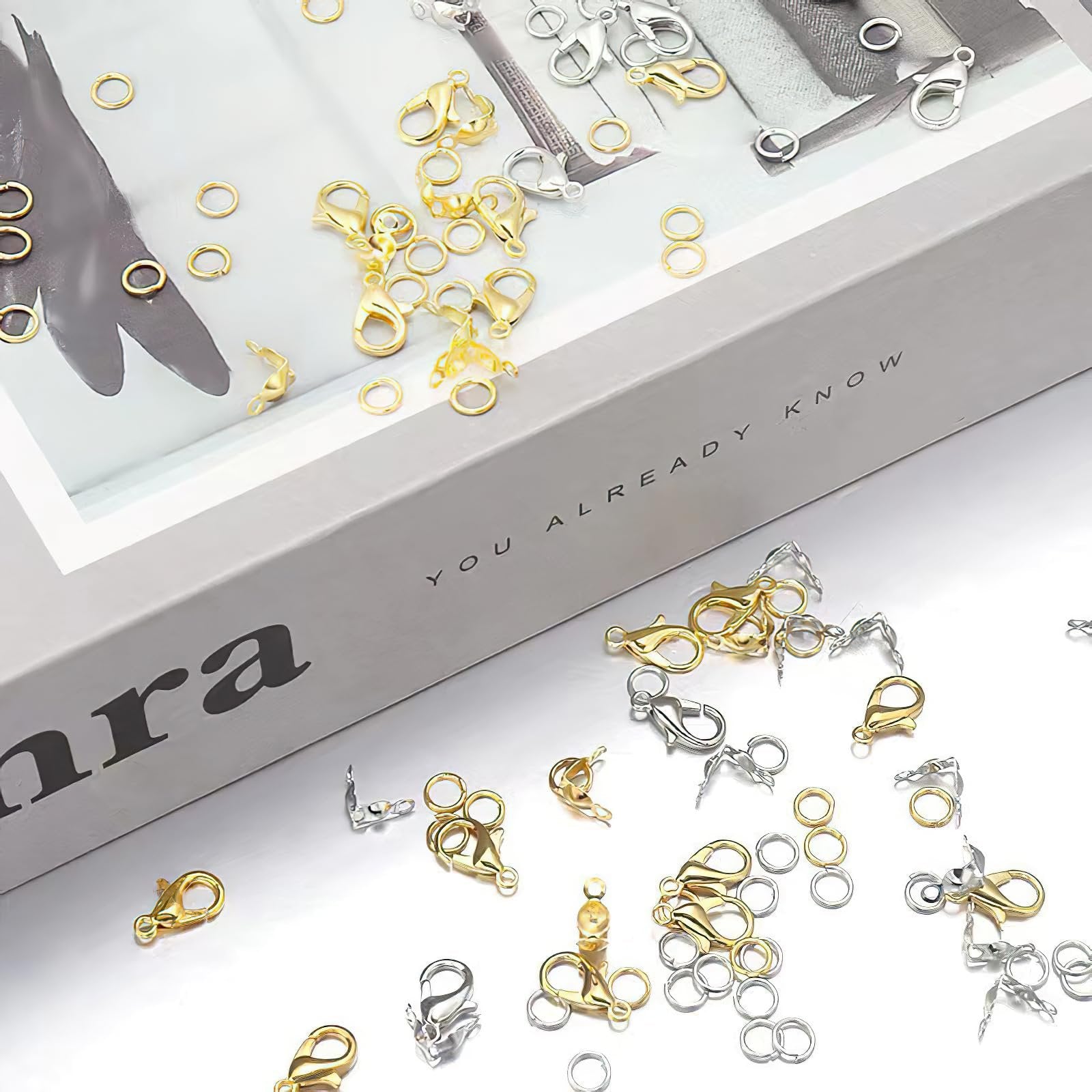 Gold and silver lobster claw clasps and jump rings scattered on a reflective surface with a book in the background.
