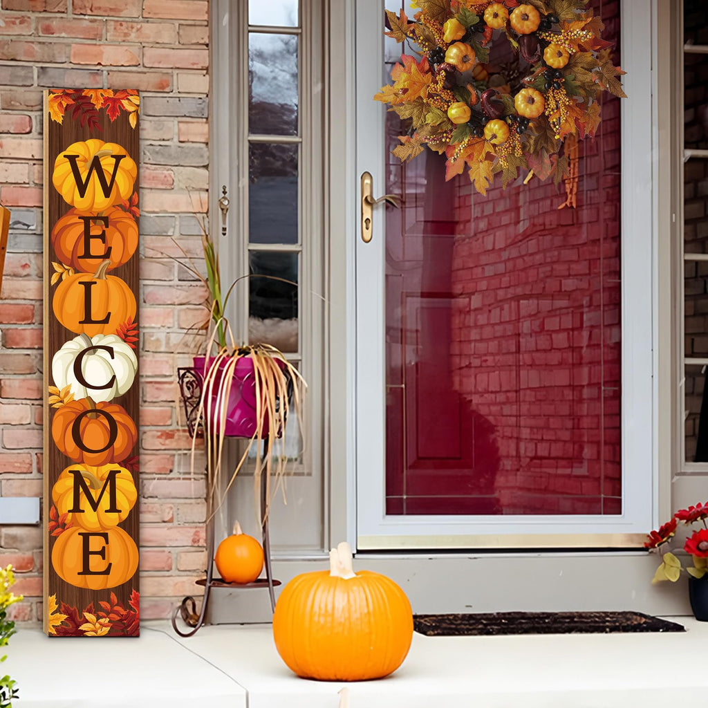 Vertical wooden porch sign with pumpkins and leaves spelling WELCOME, displayed on a porch next to a door with a fall wreath.