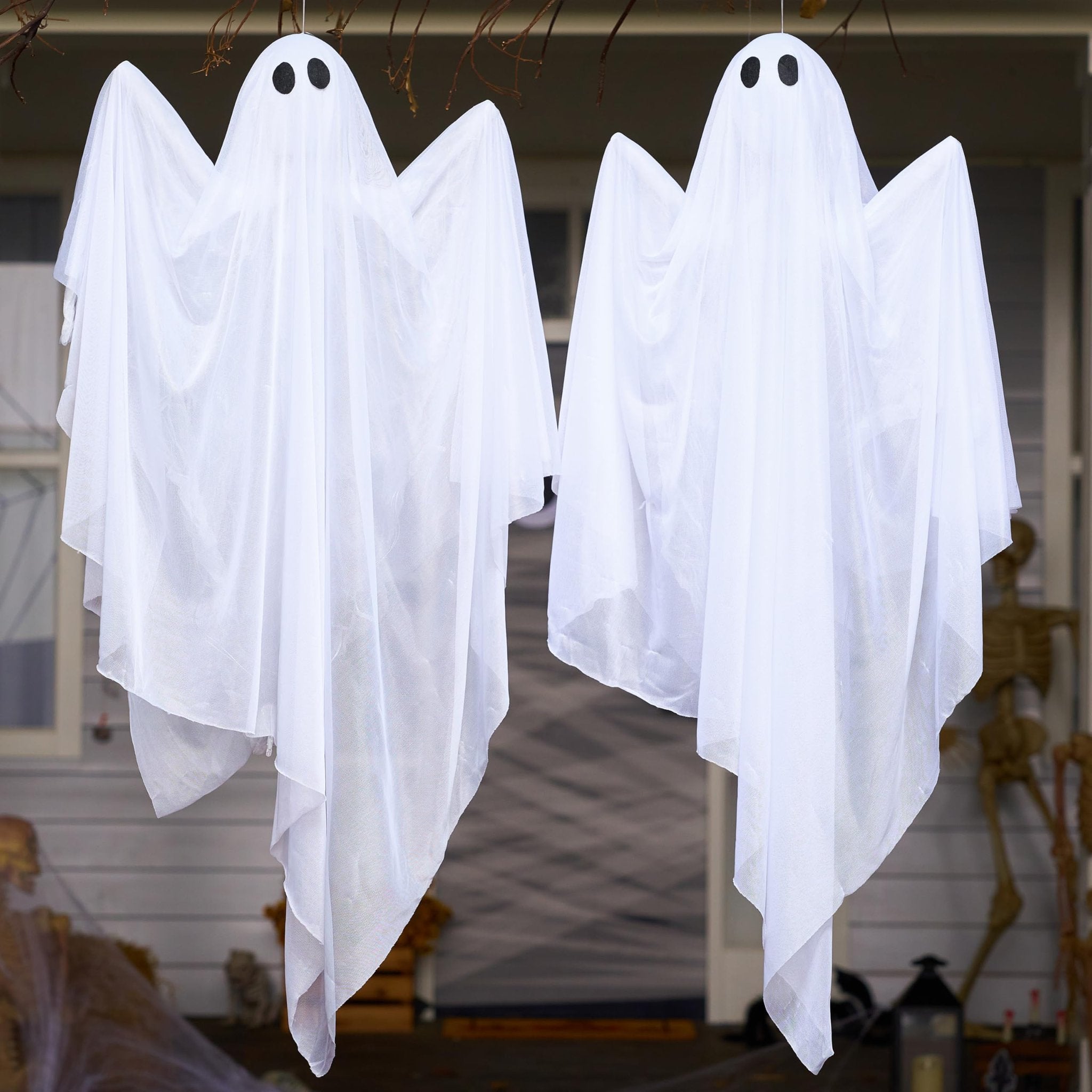 Two white hanging ghost decorations with black eyes, displayed outdoors in front of a house.