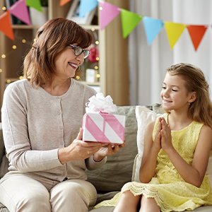 Woman giving a pink and white striped gift box with a white bow to a smiling young girl on a couch, with colorful party banners in the background.