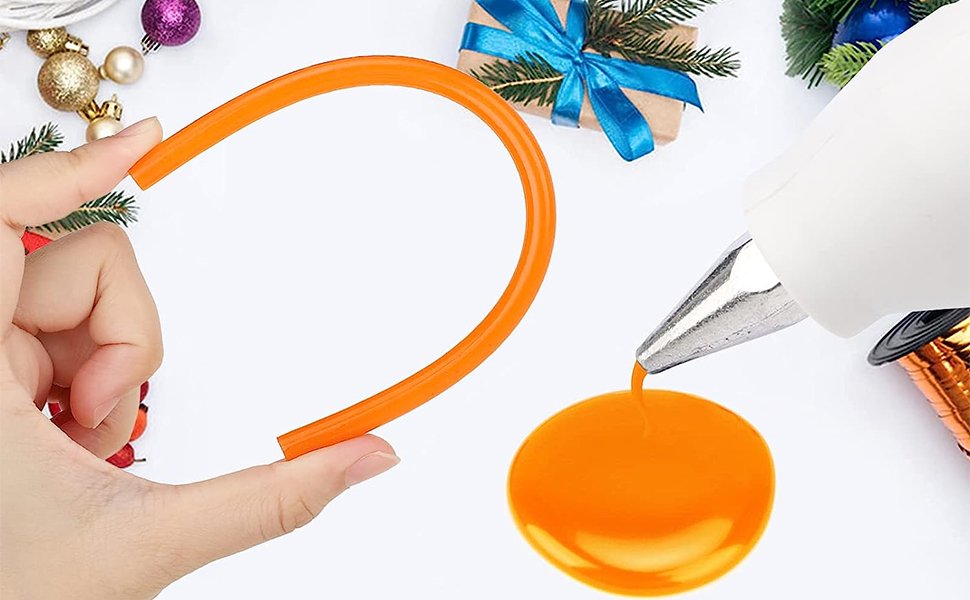 Hand holding orange mini hot glue stick with melted glue dripping from a glue gun nozzle onto a white surface with holiday decorations.