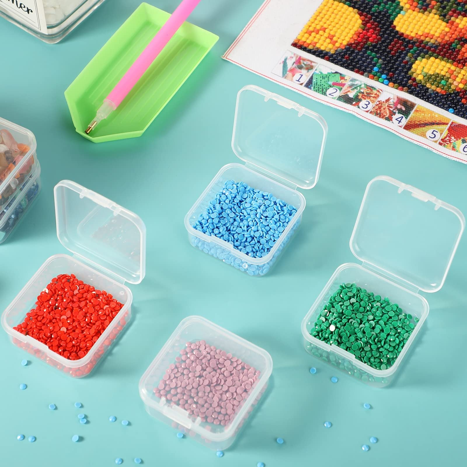 Four clear plastic storage boxes filled with red, pink, blue, and green beads, with a diamond painting kit and tool in the background.