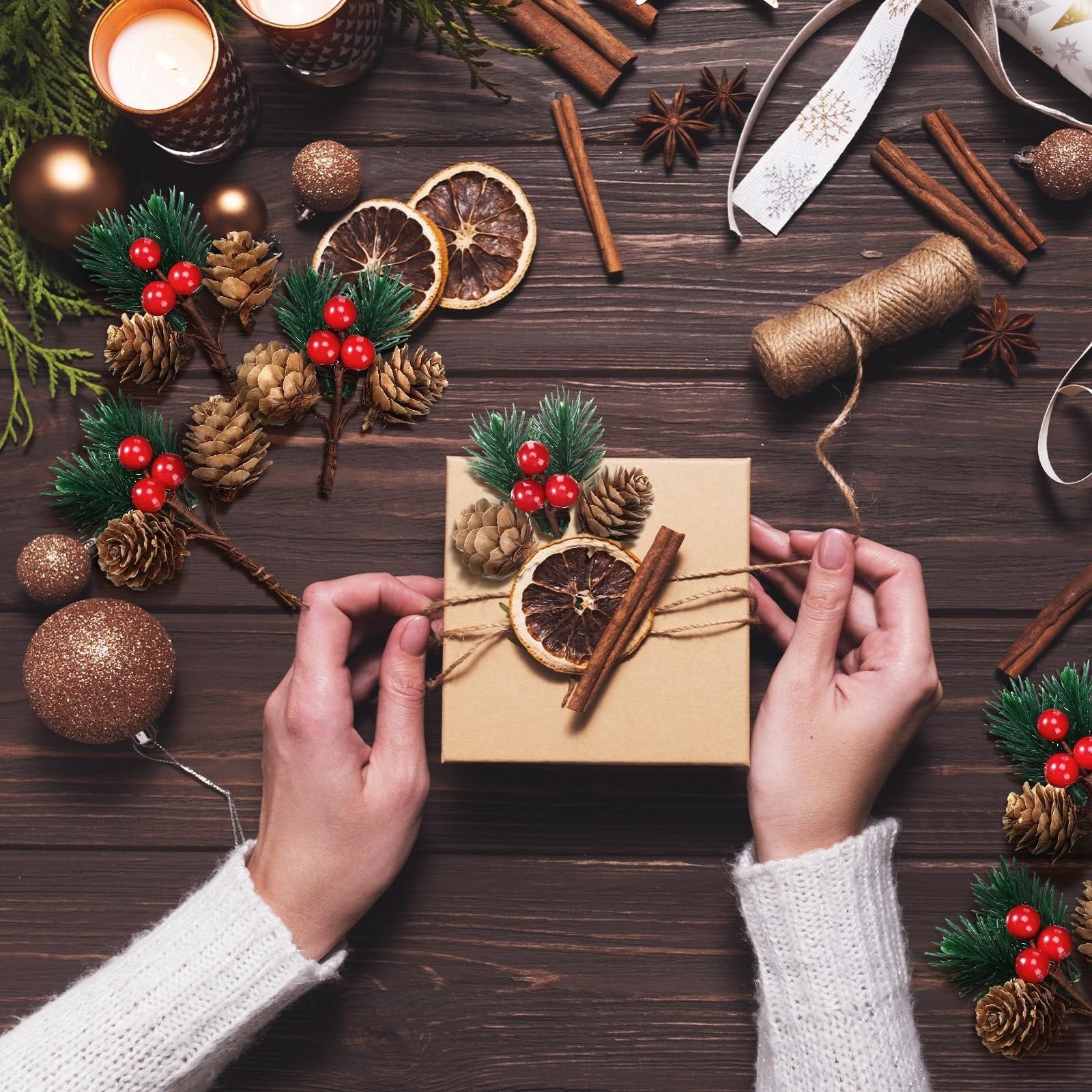 Hands tying twine around a Christmas gift box decorated with pine sprigs, pinecones, dried orange slices, and cinnamon sticks.