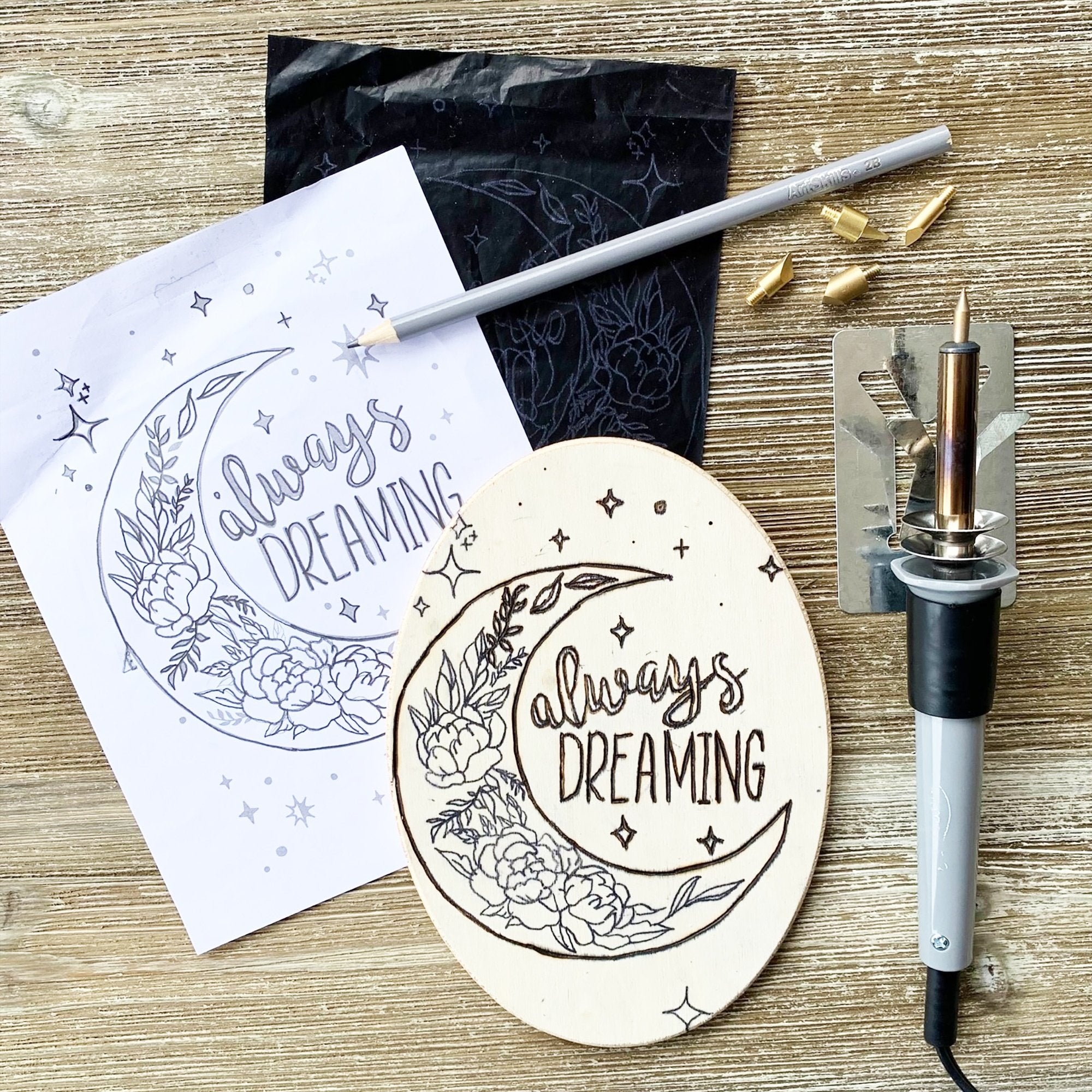 Wood burning kit components including a burner pen, tips, a wooden plaque with a moon design, and a stencil on a wooden surface.