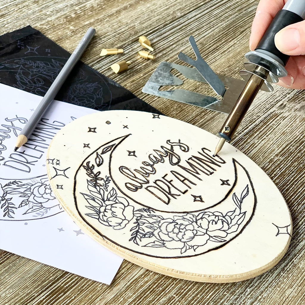 Wood burning kit in use, a hand holds a burner pen to wood plaque with moon and floral design, pencil and transfer paper nearby.