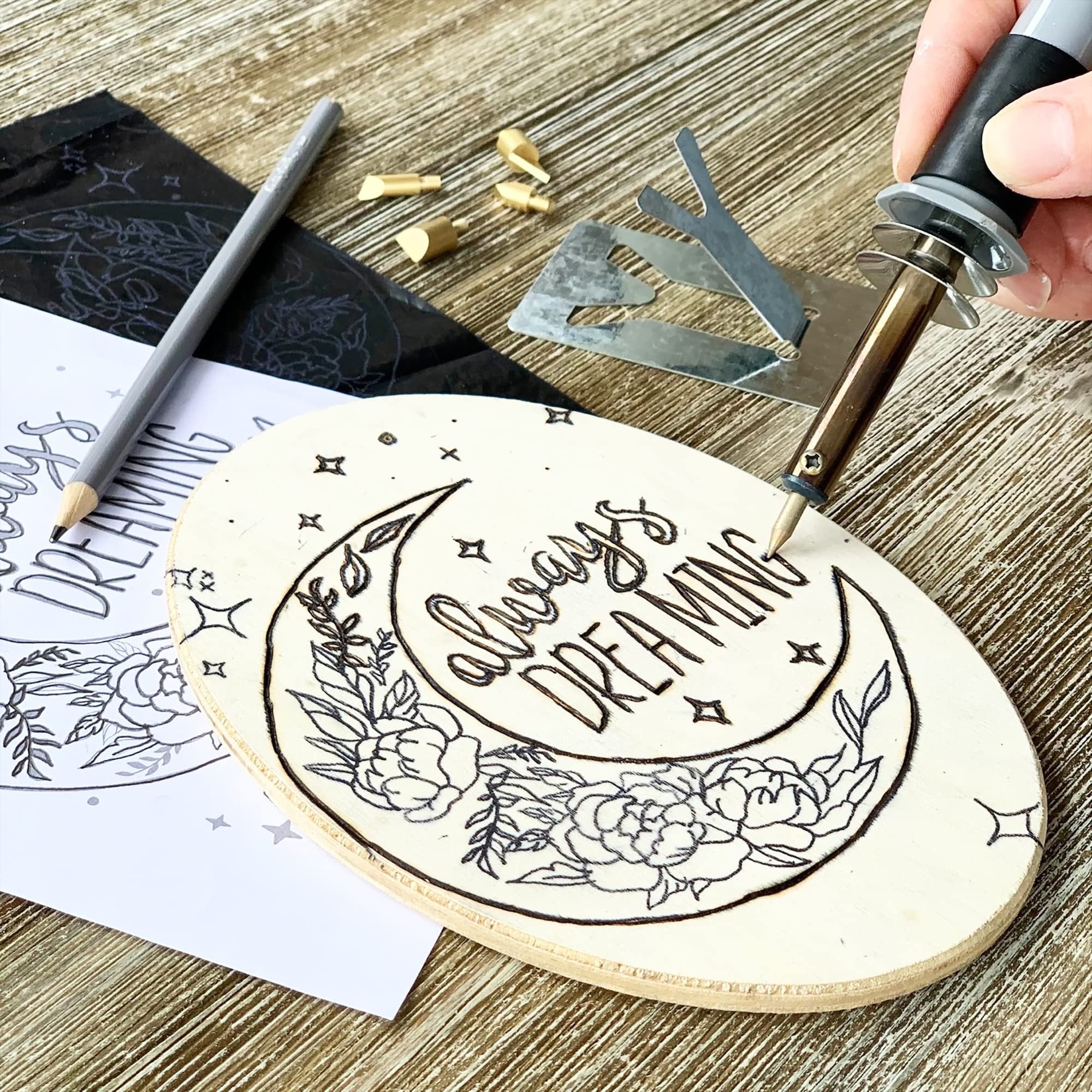 Wood burning kit in use, a hand holds a burner pen to wood plaque with moon and floral design, pencil and transfer paper nearby.
