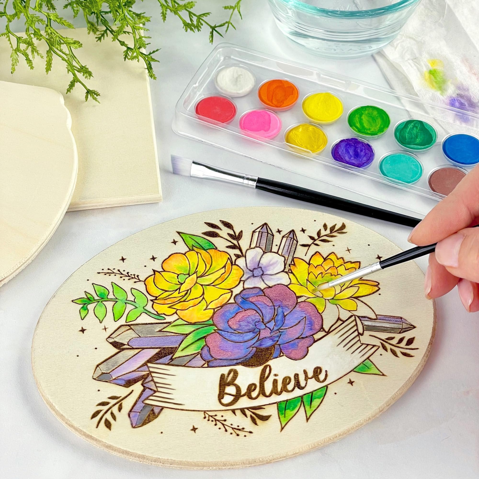 Oval wood plaque with floral and crystal design being painted by a hand holding a brush, watercolor palette and brush nearby.