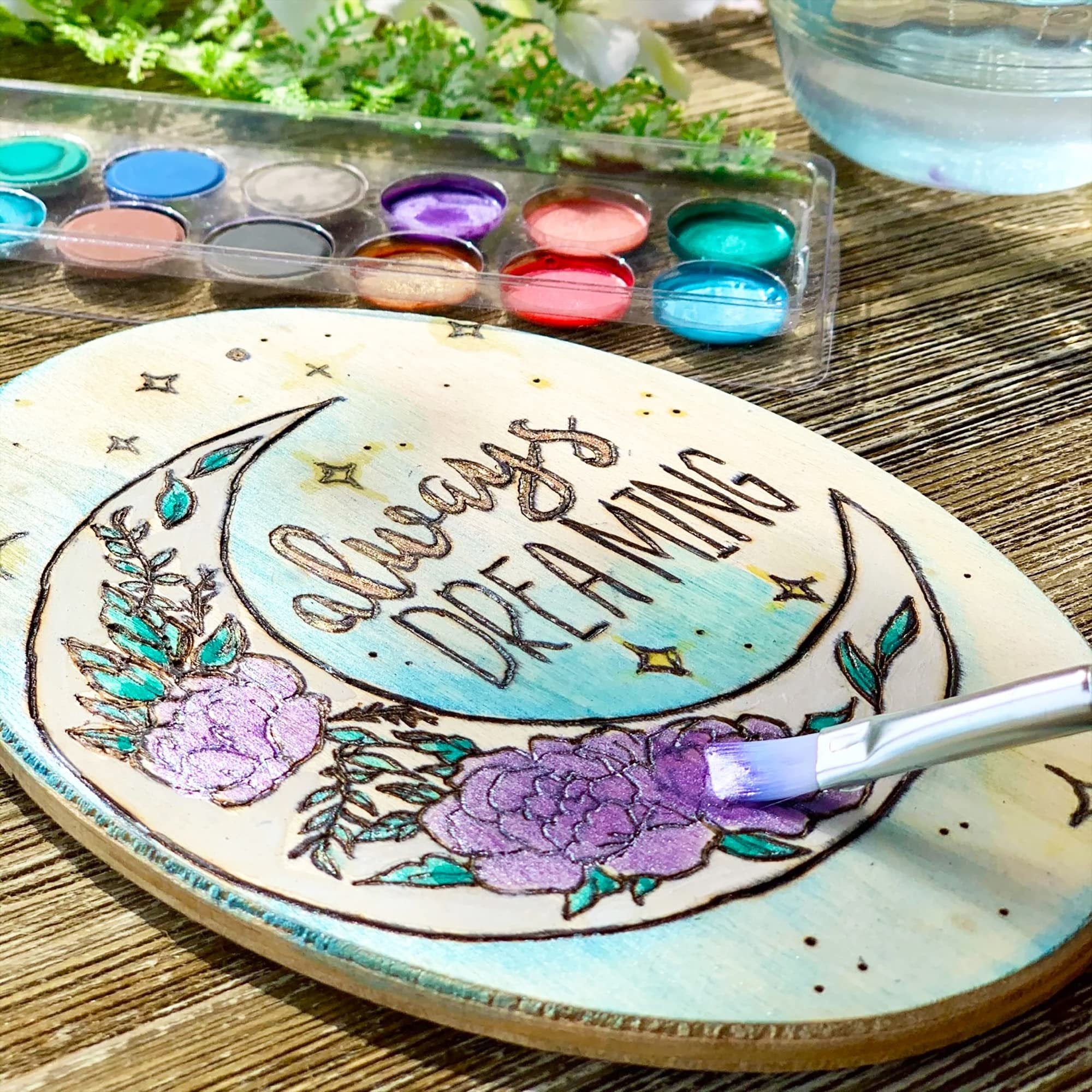 Wood plaque with moon and floral design being painted with a brush, watercolor palette in background