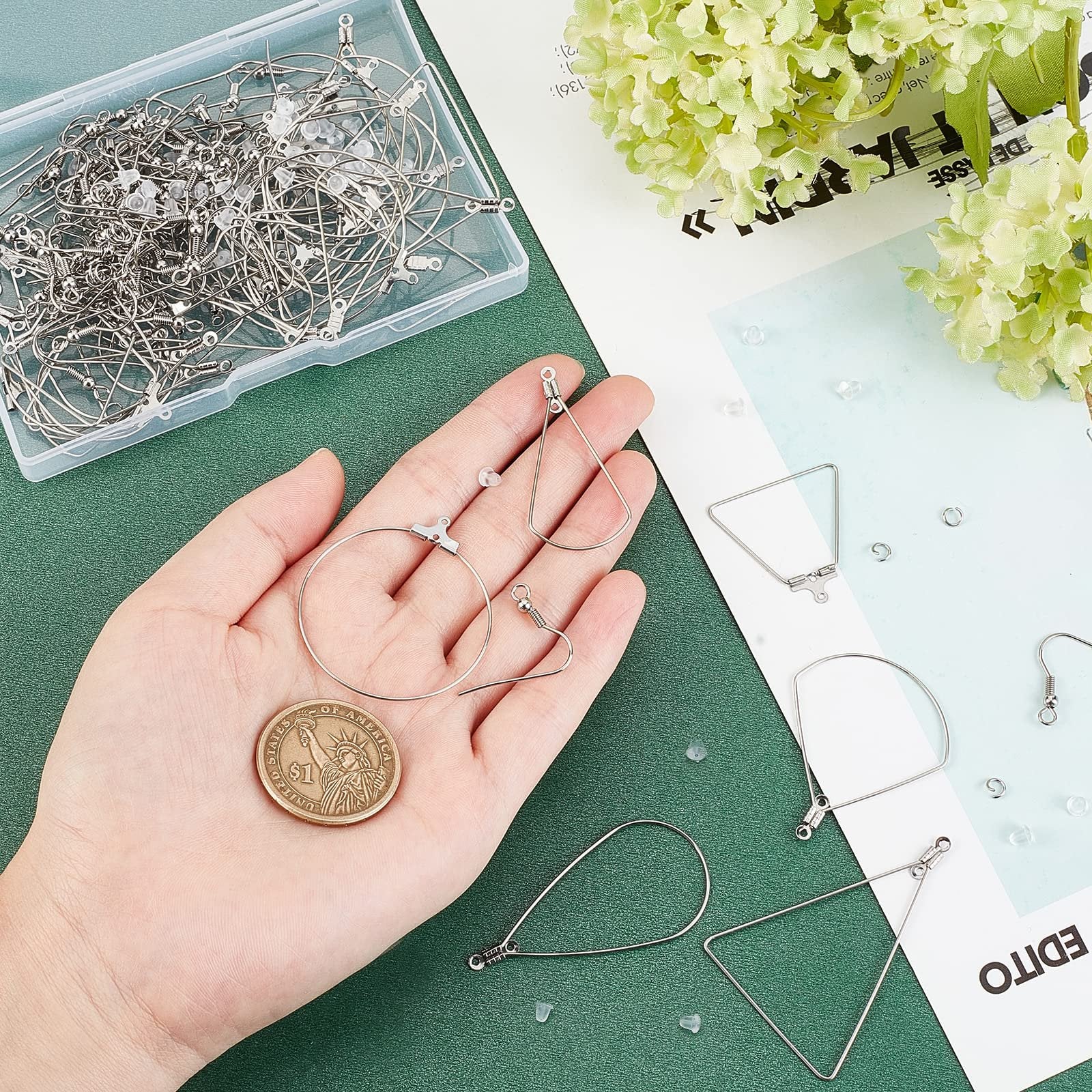 Hand holding stainless steel earring components including hoops, triangles, and hooks, with a dollar coin for scale. A clear plastic box of findings is in the background.