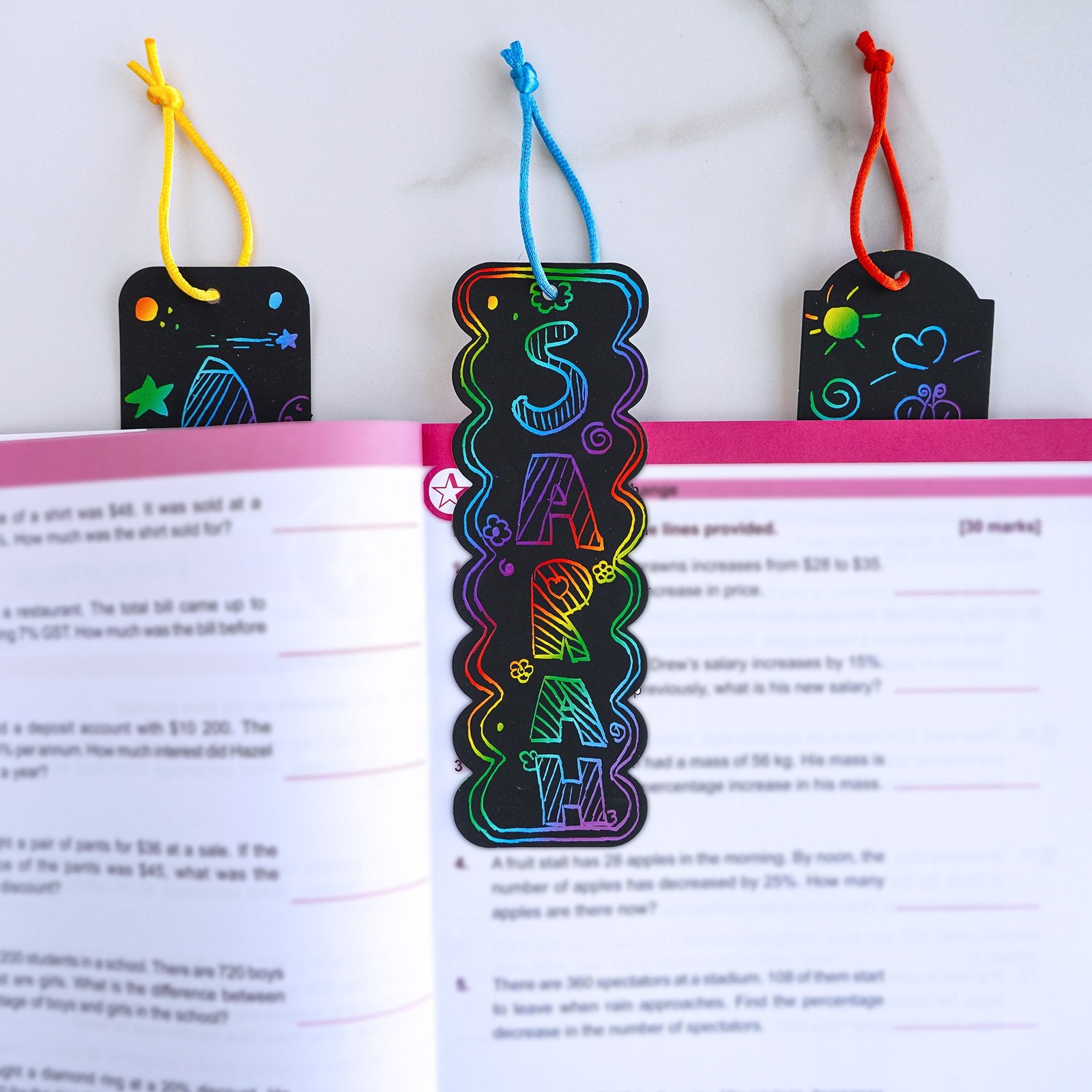Rainbow scratch art bookmarks with colorful designs and cords, resting on an open book.